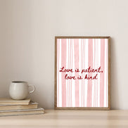 Framed print with 'Love is patient, love is kind' text on a pink and white striped background, placed on a surface with books and a mug.