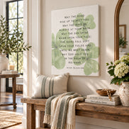 A square canvas wall art print featuring the Irish Blessing poem in black text over a white background with soft green leaf motifs, displayed in a bright, modern entryway above a wooden bench with a striped pillow and throw blanket.